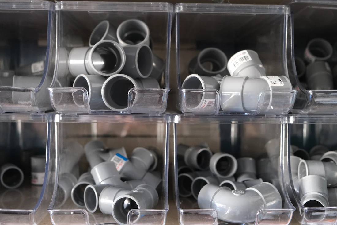 Plusieurs raccords de tuyaux PVC gris stockés dans des bacs en plastique transparent.