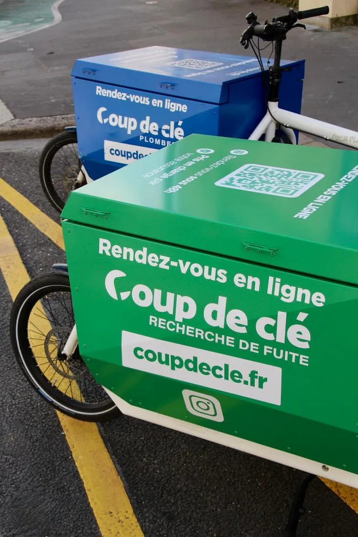Deux vélos cargos avec des caisses colorées, une verte en premier plan et une bleue à l'arrière, portant des inscriptions publicitaires pour « Coup de clé » invitant à un rendez-vous en ligne.