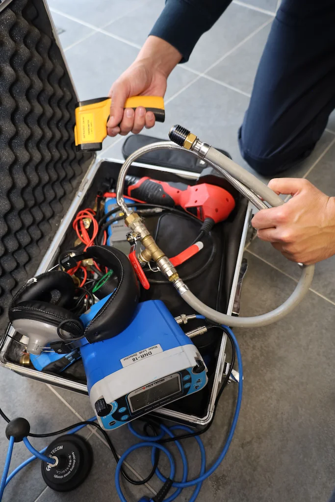 Personne tenant un détecteur de fuite jaune et un tuyau flexible au-dessus d'une mallette ouverte contenant des outils de recherche de fuite.