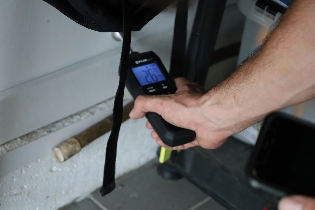 Main tenant un détecteur de fuite numérique avec un écran affichant 26.7 devant un mur blanc à côté de conduites.