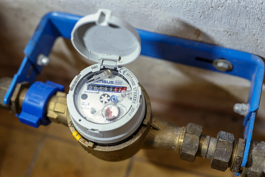 Compteur d’eau installé sur une canalisation avec un couvercle blanc ouvert et un cadran affichant des chiffres.