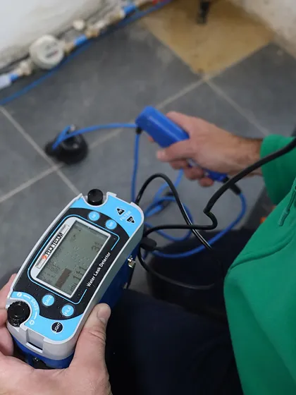 Une personne tenant un appareil de mesure bleu avec un écran et un câble bleu connecté à une sonde tenue dans l'autre main, contrôlant un dispositif au sol.