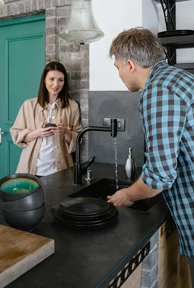 Un homme lave des assiettes dans un évier de cuisine moderne pendant qu'une femme le regarde en souriant, tenant un téléphone portable.