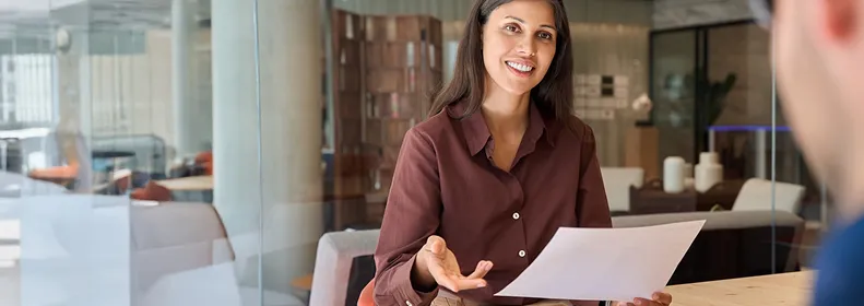 Femme souriante tenant un document, discutant avec une autre personne dans un bureau moderne avec des meubles en verre et en bois.