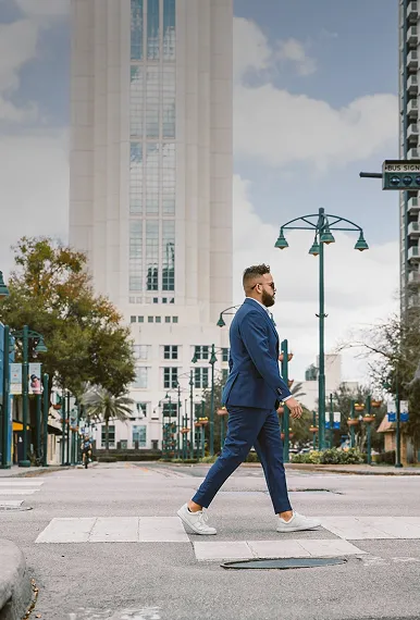 Un homme portant un costume bleu et des chaussures blanches traverse une rue en traversant un passage piéton en milieu urbain avec un grand bâtiment en arrière-plan.
