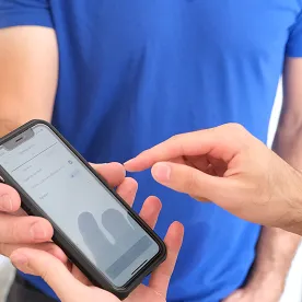 Une personne en t-shirt bleu tenant un smartphone affichant un menu, une autre main pointant l'écran.