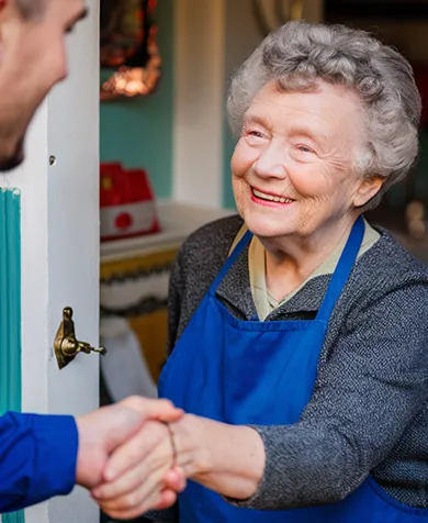 Une femme âgée souriante portant un tablier bleu serre la main d'une autre personne à la porte.