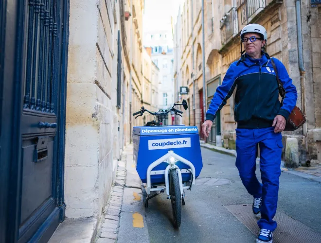 Un homme en tenue bleue avec casque marche à côté d'un vélo cargo bleu dans une rue étroite en pierre, avec un panneau indiquant dépannage en plomberie et coupedele.fr.
