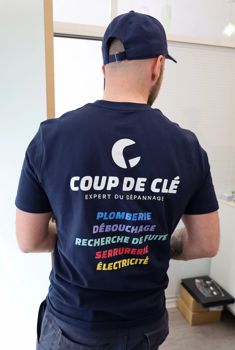 Homme vu de dos portant un t-shirt bleu marine avec le texte Coup de Clé expert du dépannage et les services plomberie, débouchage, recherche de fuite, serrurerie, électricité.