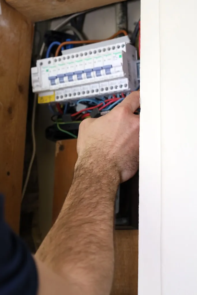 Main tenant la porte d'un panneau électrique avec disjoncteurs et câbles visibles.