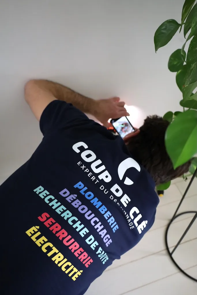 Homme en polo bleu foncé avec texte de services plomberie, électricité, serrurerie sur le dos, travaillant au mur.