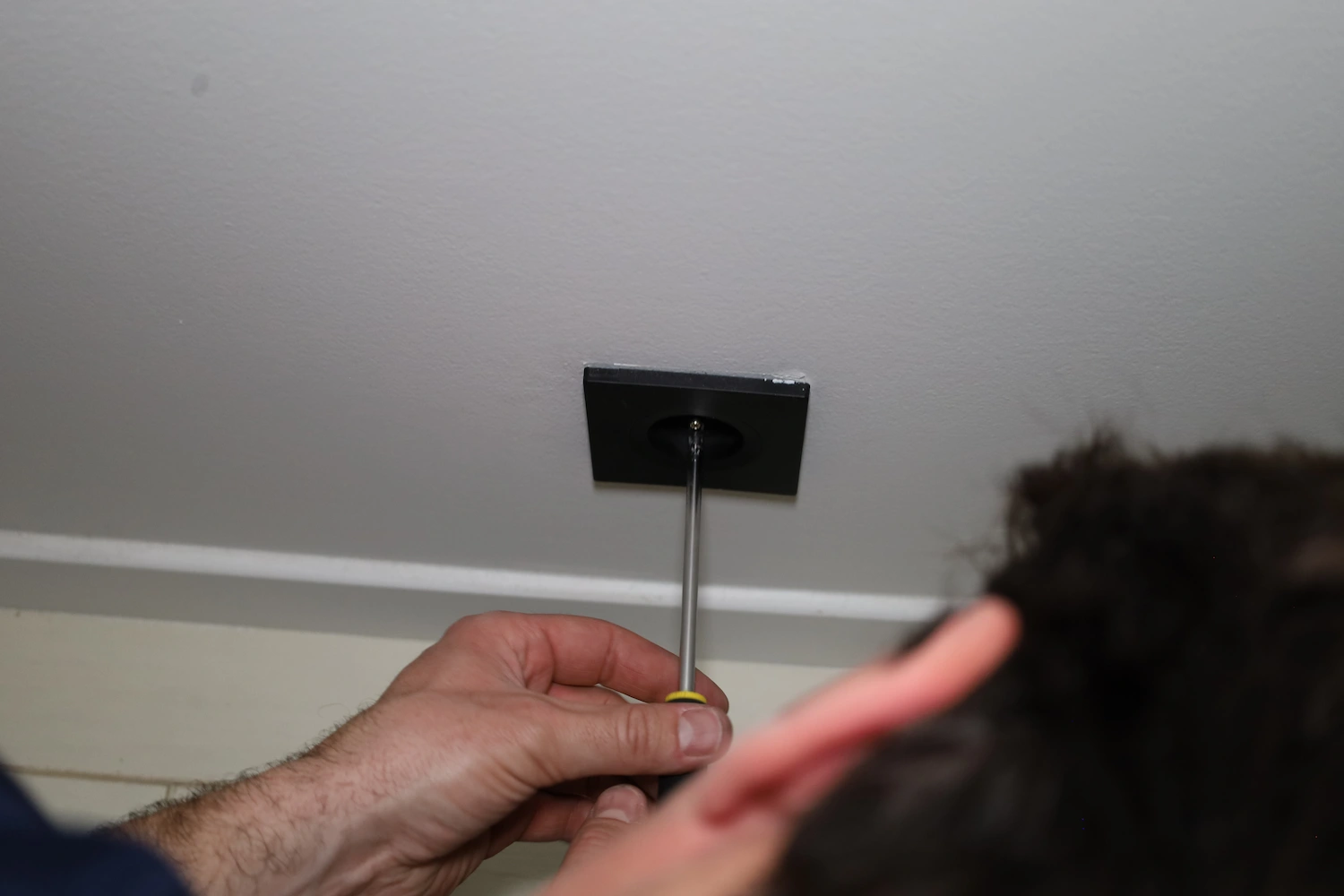 Une main utilise un tournevis pour réparer une prise électrique au plafond.