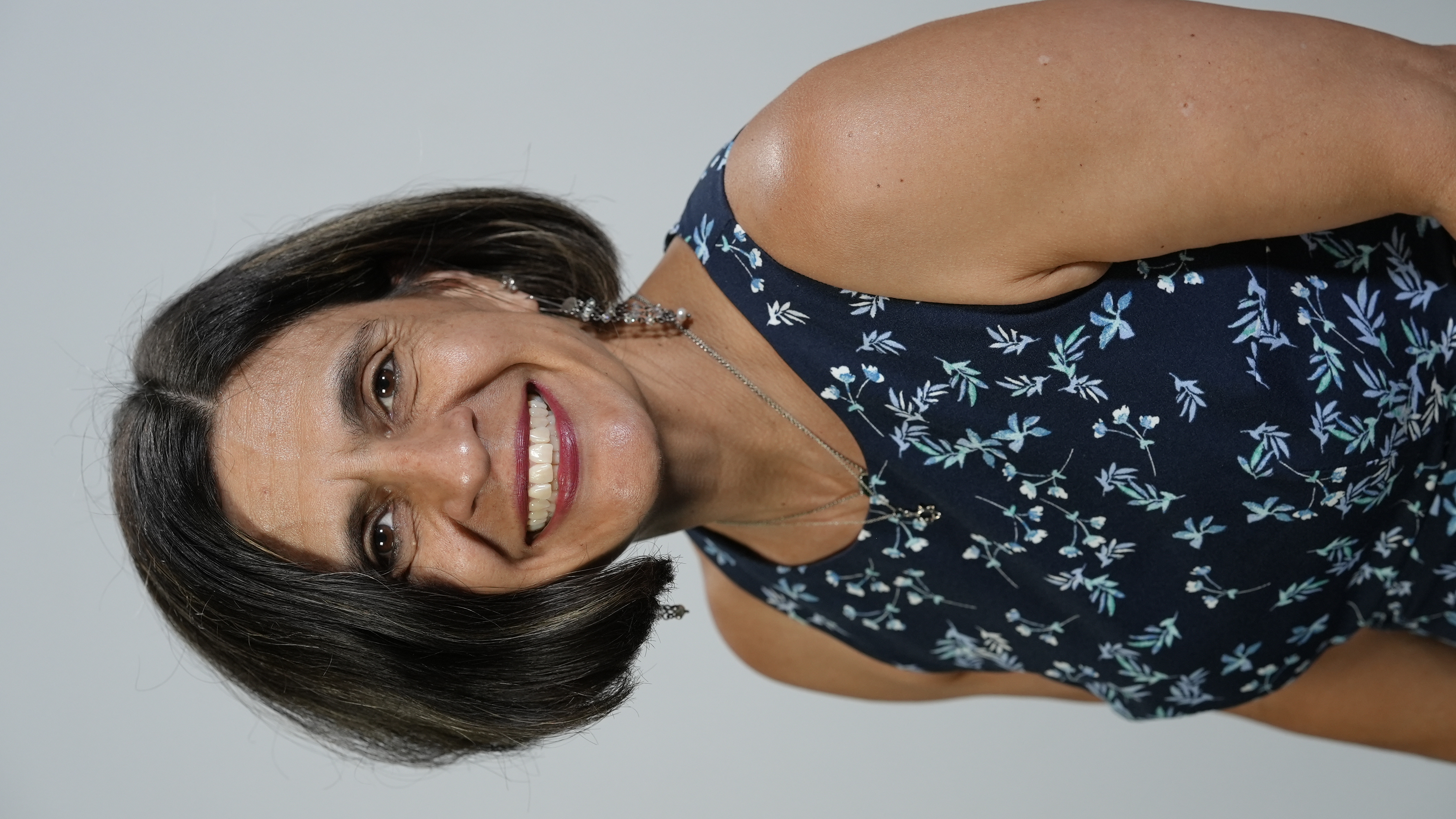 A woman wears a big smile with a short bob, dangly earrings, and a navy blue tank top with blue and white flowers. 