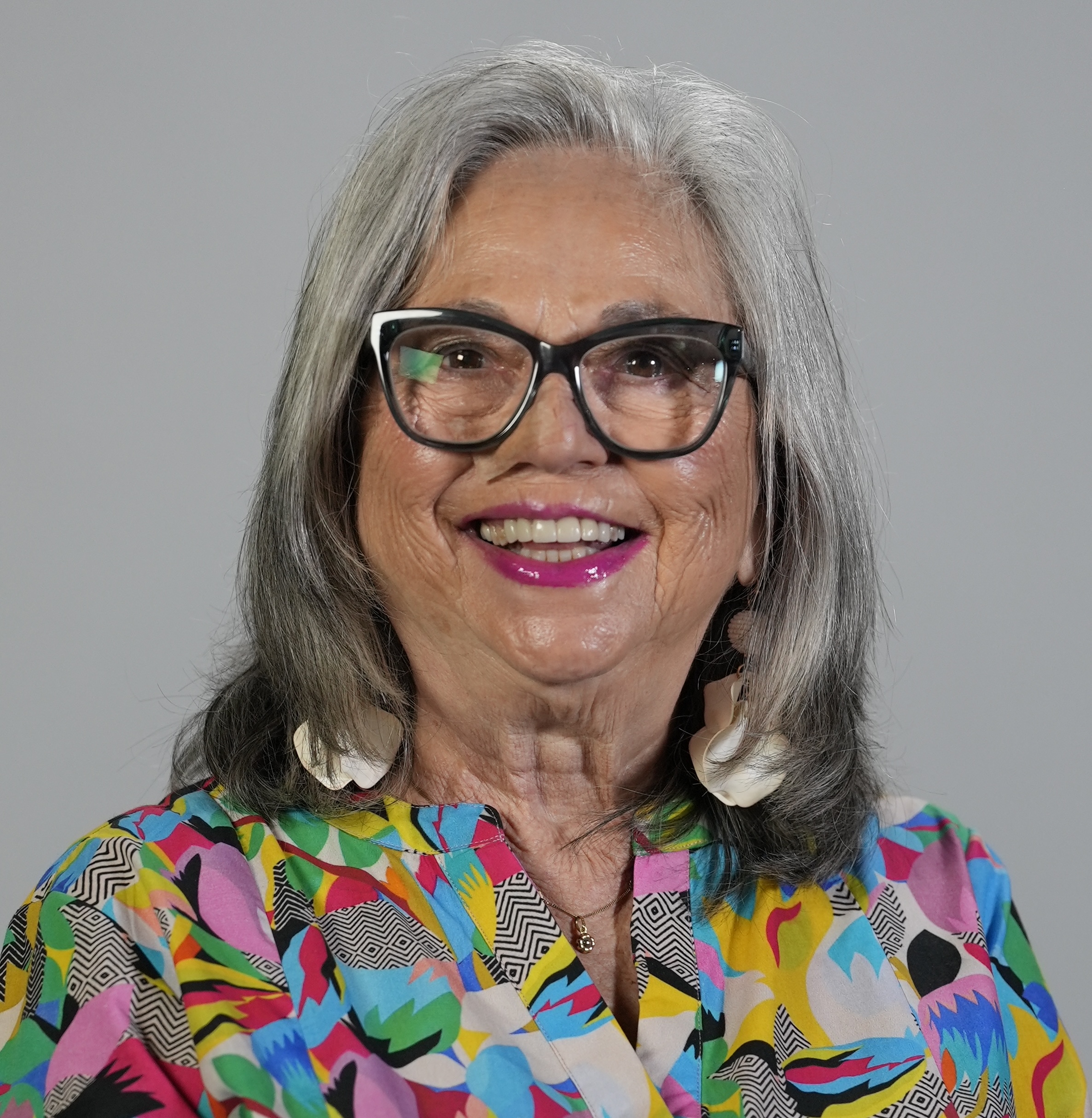 Ana wears a rainbow patterned shirt with white statement earrings. She wears black glasses with large frames and fun pink lipstick