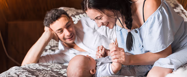 Parents smiling and playing with their baby lying on a bed with patterned bedding.