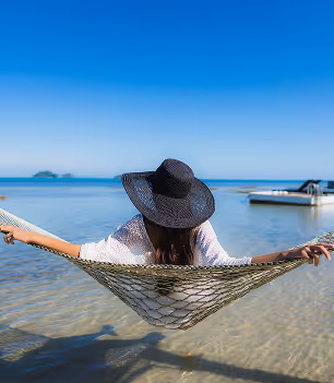 Person wearing a black hat relaxing in a hammock over shallow clear water with a boat in the background under a blue sky.