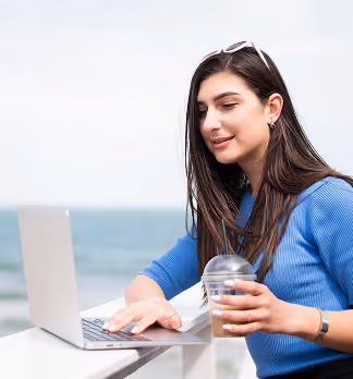 Woman in blue sweater working on a laptop outdoors by the sea, holding a drink cup.