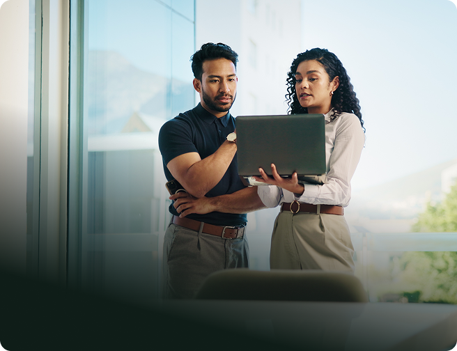 Man and woman standing near window looking at laptop screen together in an office setting.