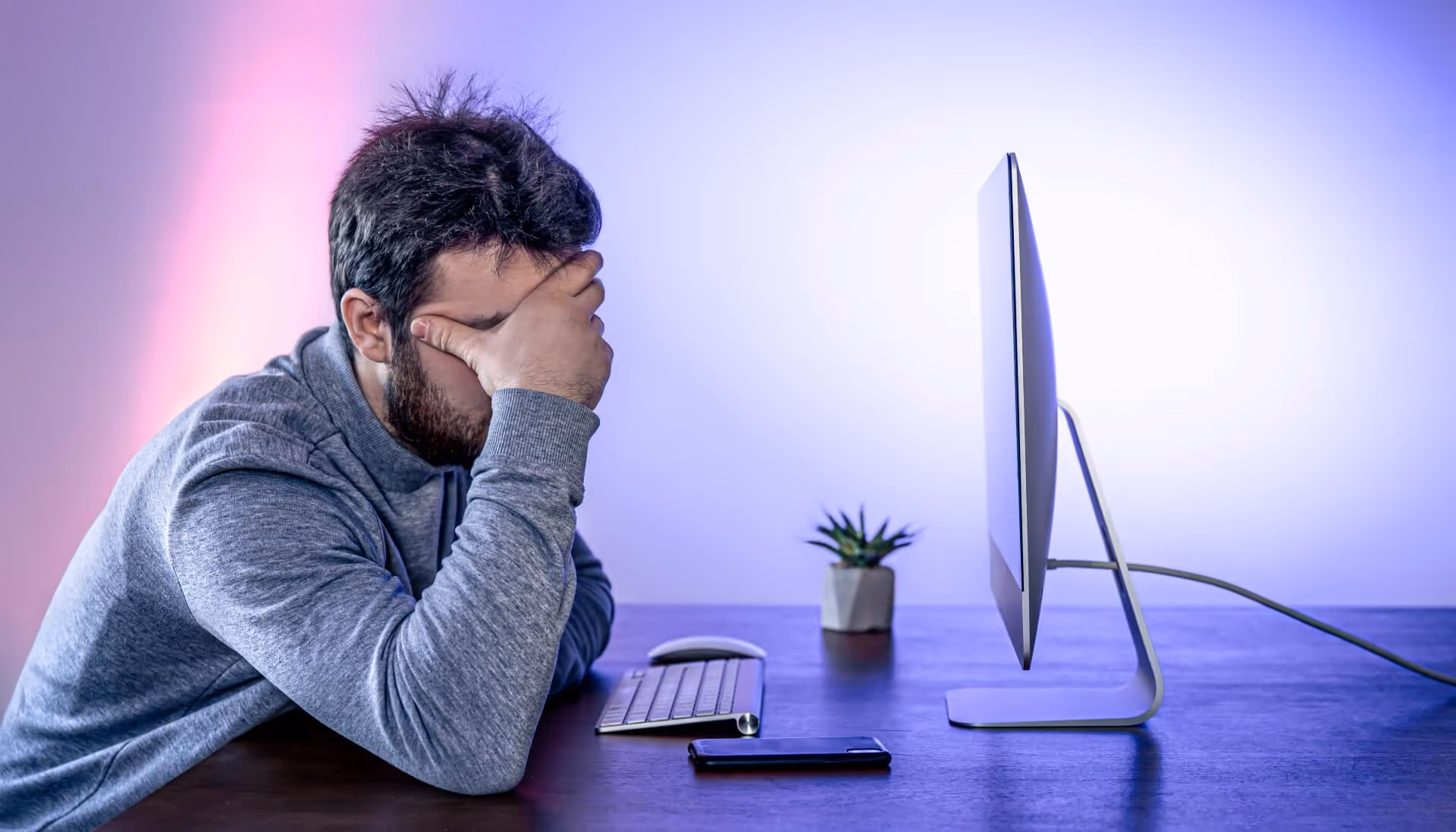 A tired man sits in front of a computer covering his face with his hand