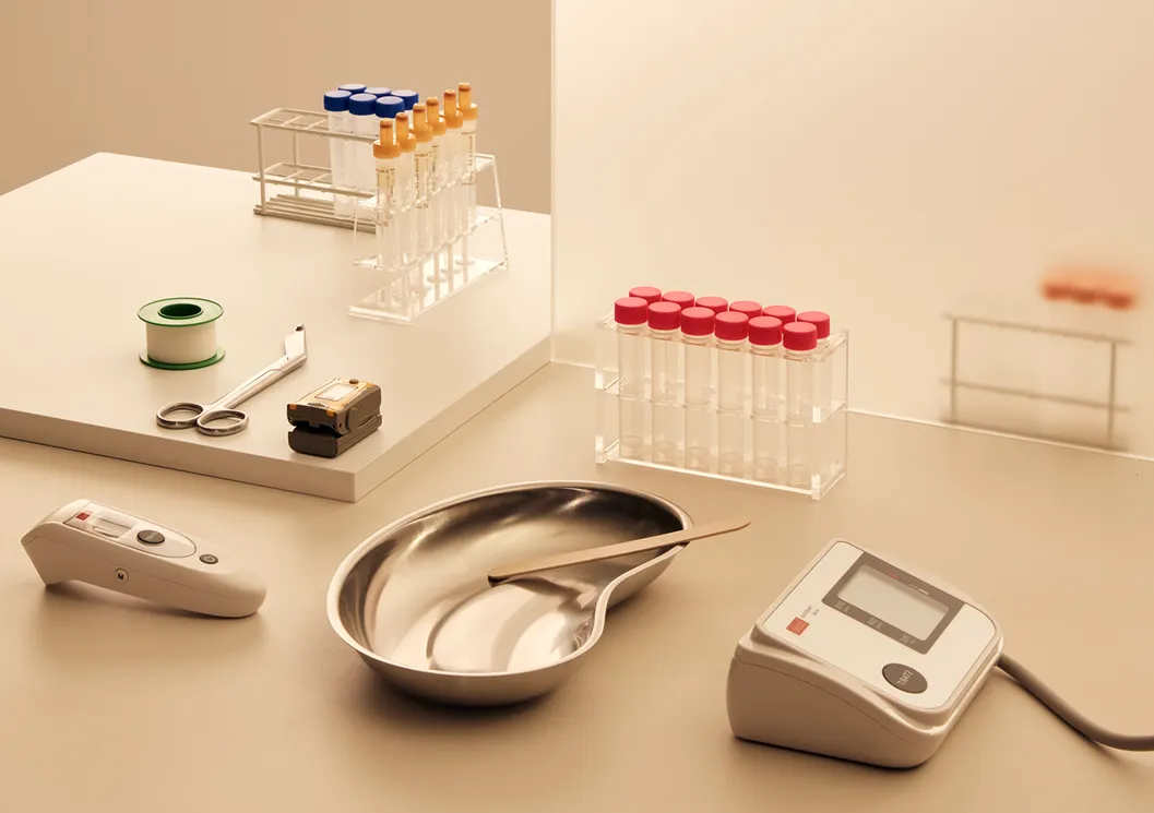 Ein medizinischer Arbeitsplatz mit verschiedenen Laborutensilien, darunter Reagenzgläser in Gestellen (mit roten, blauen und gelben Deckeln), eine Metallschale mit Pinzette, ein digitales Blutdruckmessgerät, ein Thermometer, eine Schere, medizinisches Klebeband und ein Abroller. Die Szene ist in warmem Licht gehalten und wirkt ordentlich und steril.
