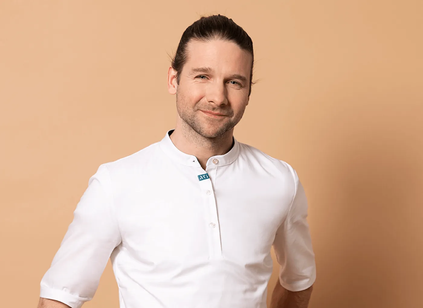 Ein lächelnder Mann mit braunen Haaren, zusammengebunden, trägt ein weißes Hemd vor einem beigen Hintergrund.