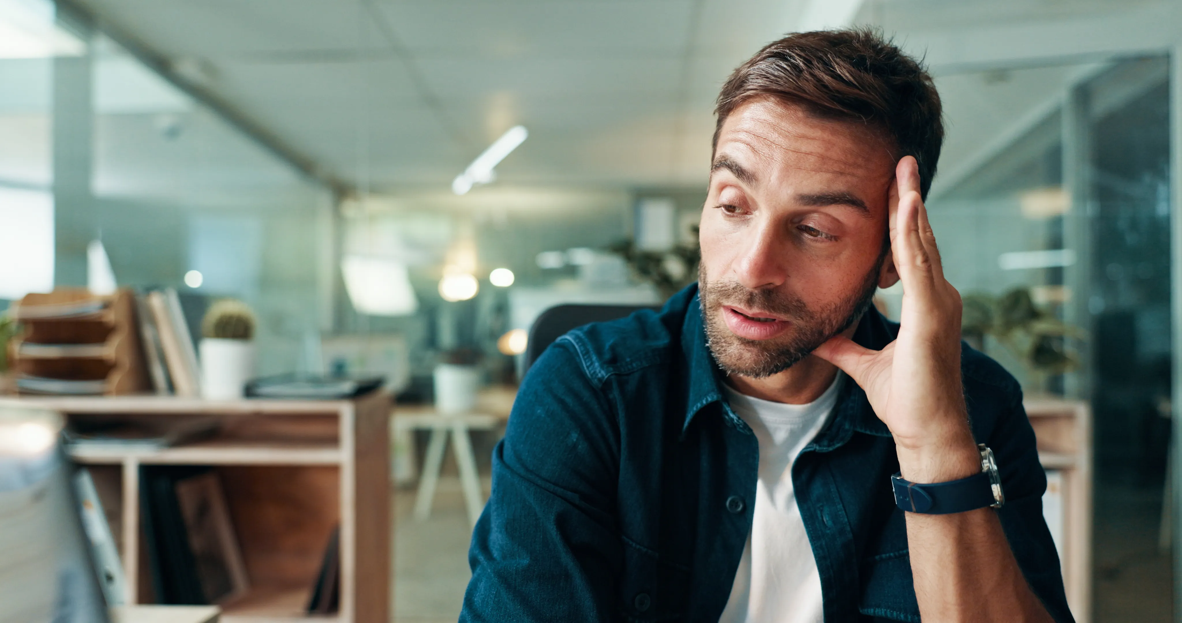Mann mit Bart und blauer Uhr sitzt nachdenklich am Schreibtisch im modernen Büro.