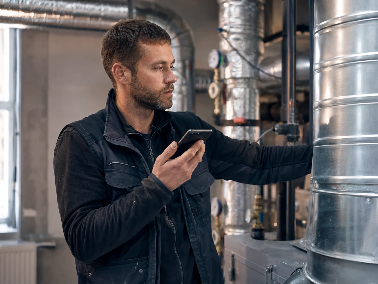 Haustechniker im Technikraum prüft mit dem Smartphone eine Lüftungsanlage