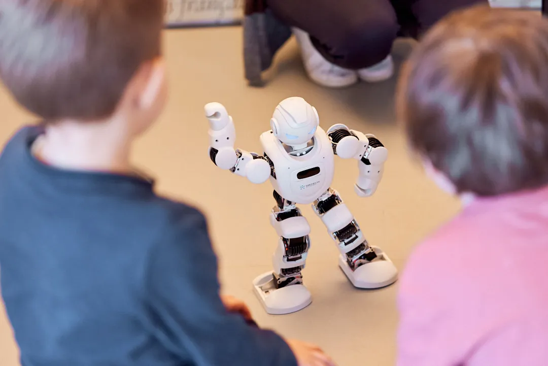 Zwei Kinder beobachten einen interaktiven, weißen, menschenähnlichen Roboter, der auf dem Boden spielt. Der Roboter hat einen Arm gehoben und scheint sich mit ihnen zu unterhalten. Im Hintergrund sind die Beine eines Erwachsenen zu sehen, der die Interaktion beobachtet.