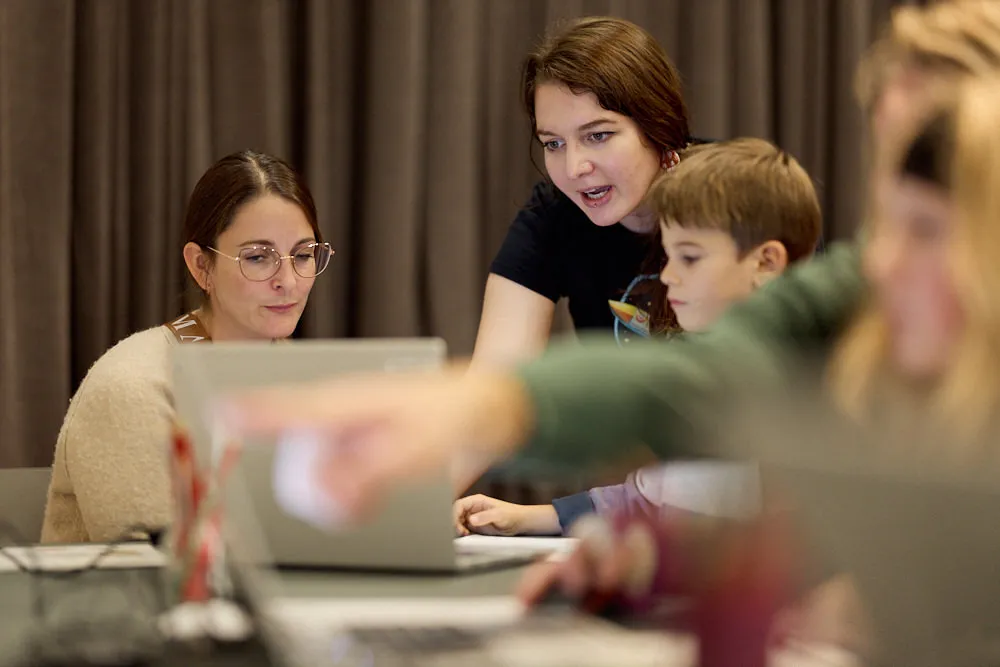 Eine Gruppe von Menschen sitzt an Tischen in einem modernen Raum und ist mit Laptops beschäftigt. Es sind mehrere Kinder und Erwachsene anwesend.