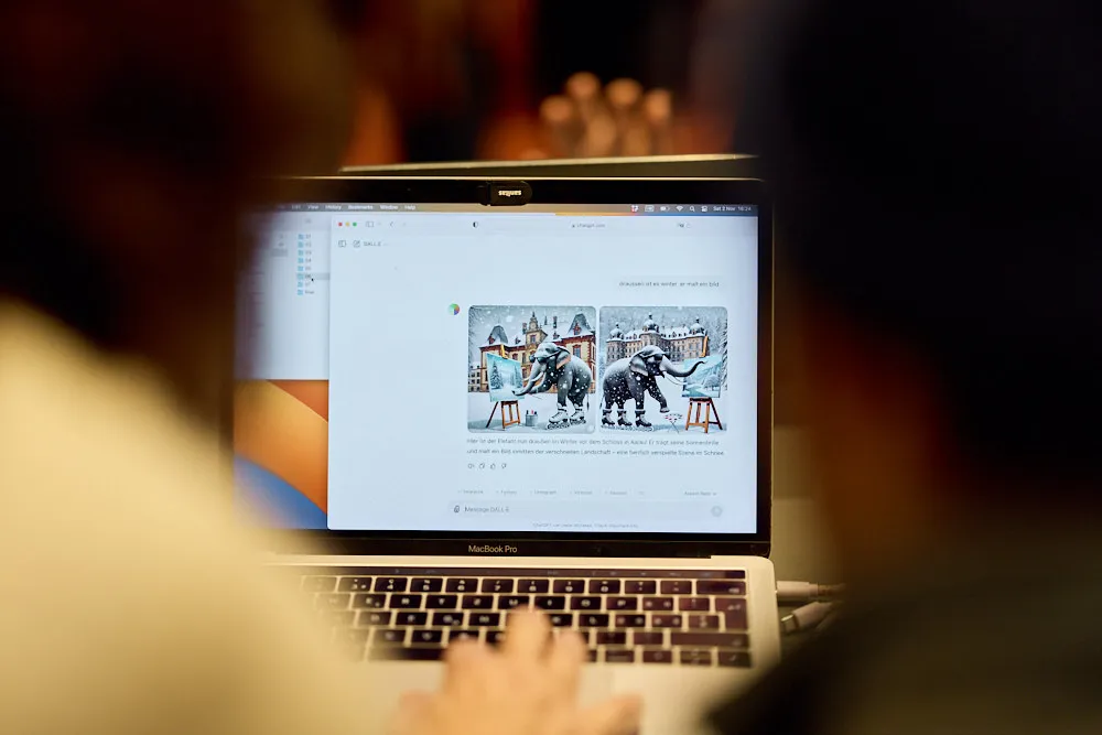 Zoom auf Laptop-Screen: Darauf sind zwei Bilder nebeneinander zu sehen von einer ähnlichen Szene, aber leicht anders umgesetzt. Es ist jeweils ein Elefant zu sehen auf Schlittschuhen, der auf einer Staffelei ein Bild malt. Im Hintergrund ist ein grosses Stadthaus zu sehen. Beide Bilder scheinen KI-generiert zu sein.