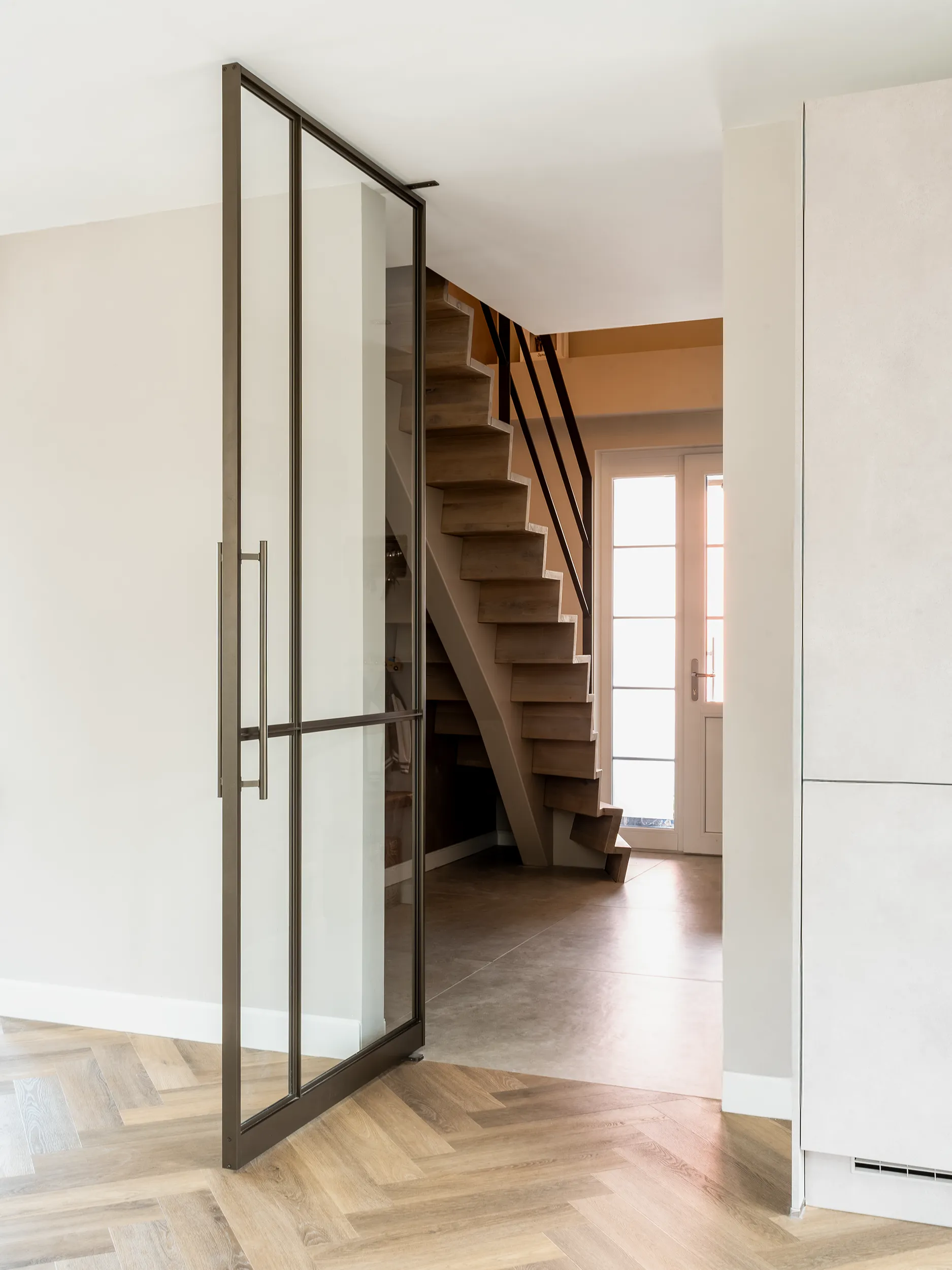Geöffnete Aluminium-Pivot-Lofttür mit Glas und schmalem T-Profil-Rahmen in Bronze, hoher Griff rechts, dahinter Holztreppe mit schwarzem Geländer und Glastür zum Garten, Boden mit Fischgrät-Parkett.