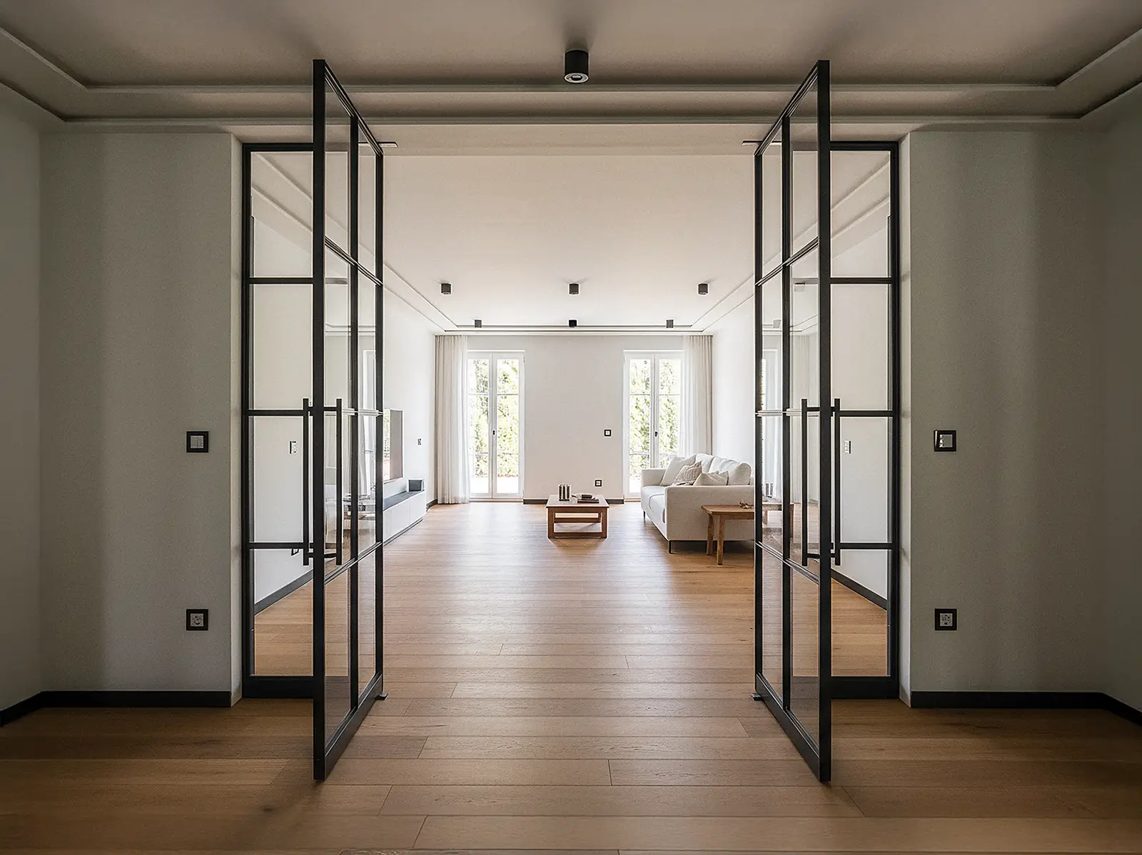 Blick in ein modernes Wohnzimmer mit zwei geöffneten Pivot-Lofttüren aus schwarzem Aluminium mit Glas und schmalen Sprossen. Die Türen sind mittig angeordnet und rechts und links von festen gläsernen Seitenelementen eingerahmt. Dahinter liegt ein heller Raum mit Eichenparkett, einem hellen Sofa und bodentiefen Fenstern, durch die Tageslicht hereinfällt.