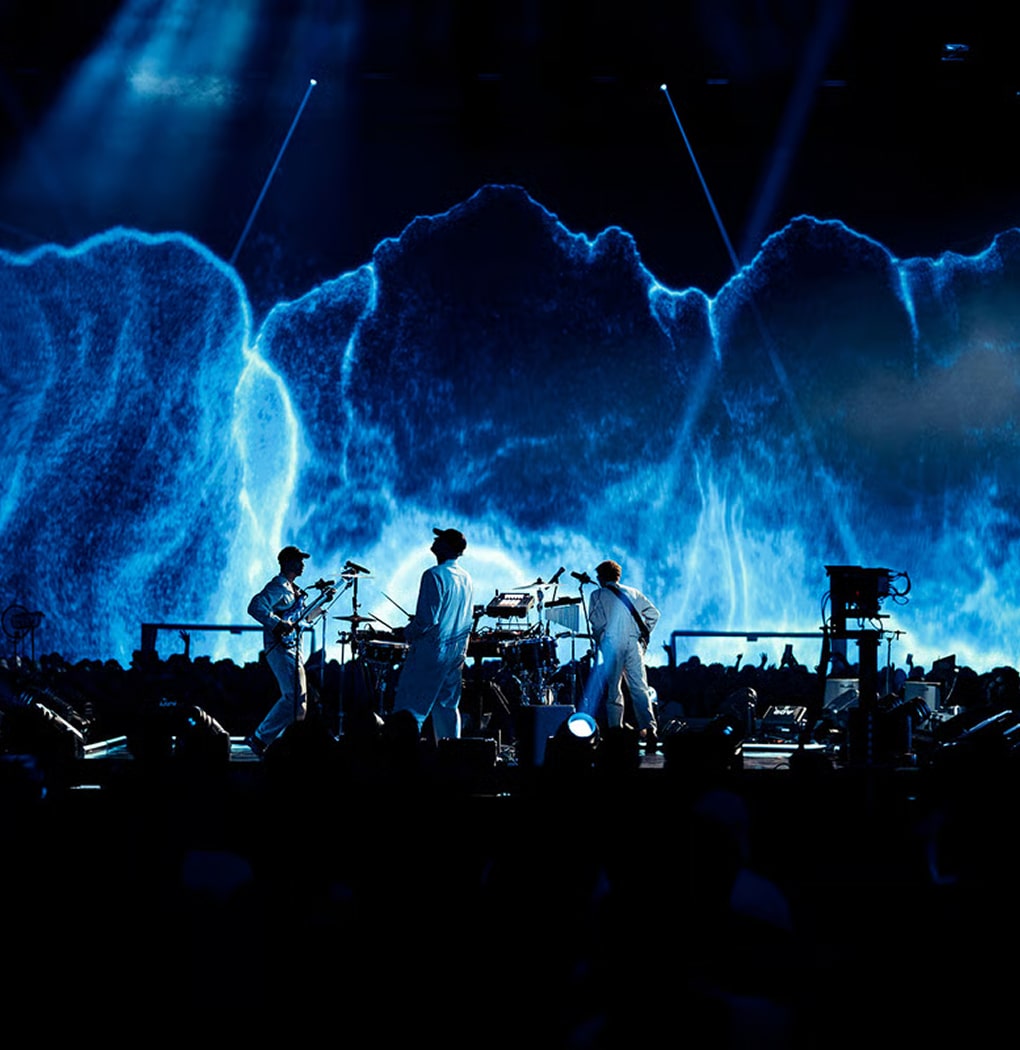 Three musicians in white outfits performing on stage with a large blue abstract digital backdrop and an audience in front.