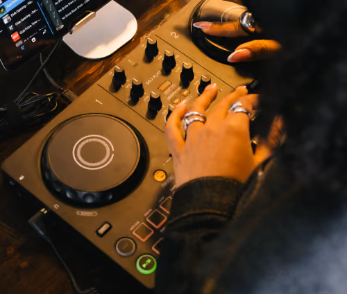 Person adjusting knobs on a DJ controller with headphones and a monitor in the background.