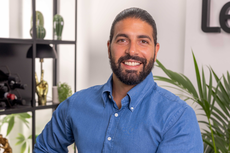 Image of Christopher Adamou, CEO and Founder of Lendhub, sat wearing a blue button-up shirt, seated in a modern office with plants and shelves in the background.
