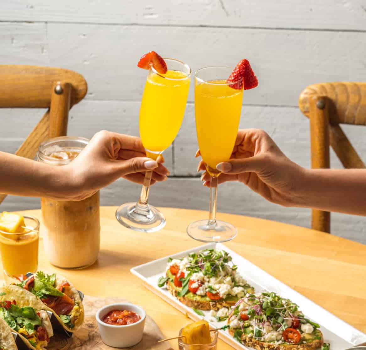Two hands clinking glasses of orange mimosa garnished with strawberry slices over a table with tacos, salsa, avocado toast, and iced coffee.