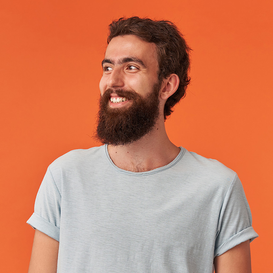 Smiling young man with a full beard and curly hair wearing a light gray t-shirt against an orange background.