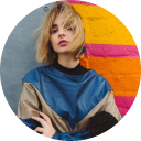 Portrait of a woman with short blonde hair wearing a stylish blue and beige top standing against a colorful wall.