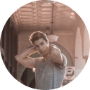 Young man with tousled hair wearing a sleeveless grey shirt posing with his hands behind his head.