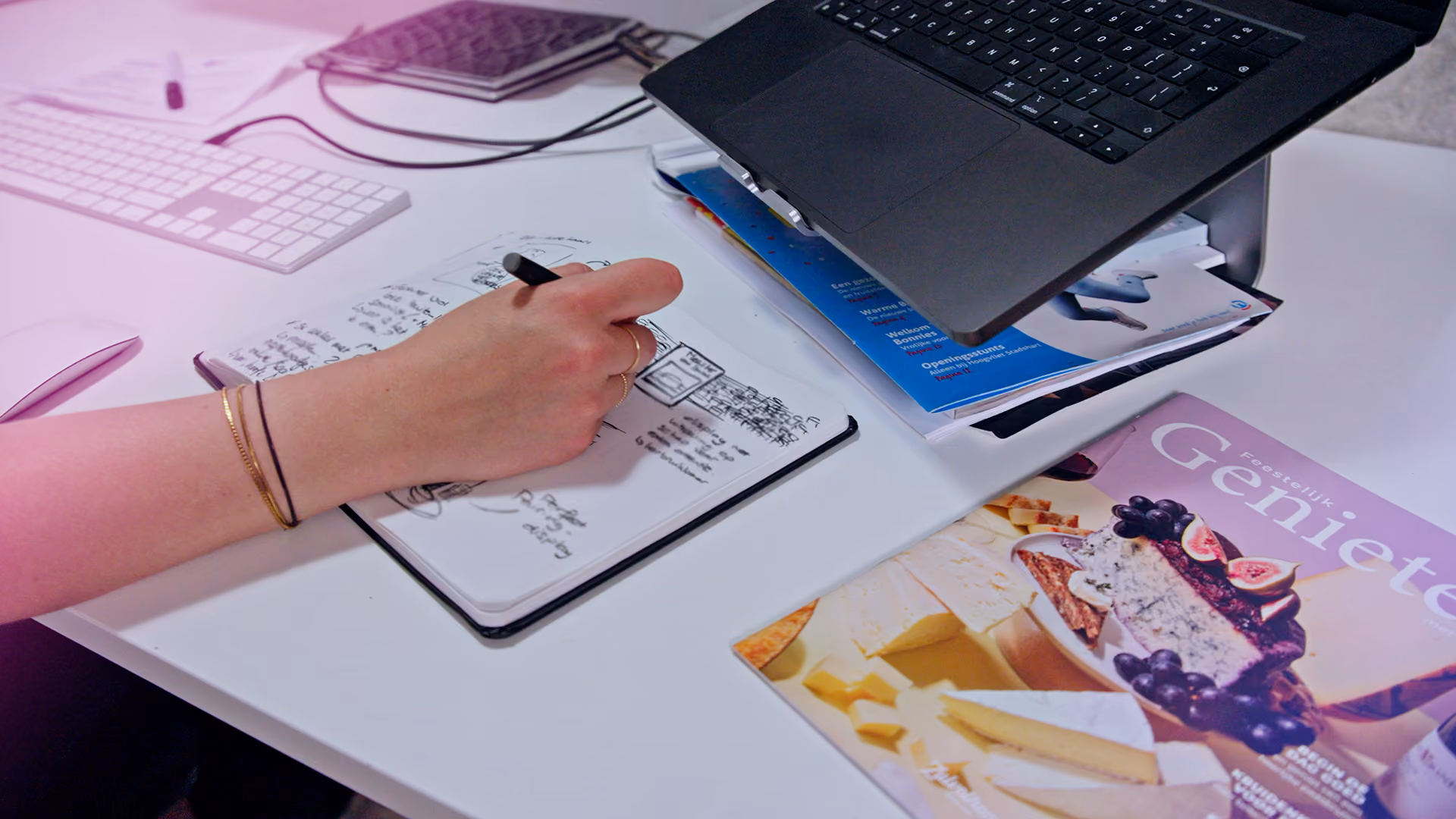 Hand schrijft aantekeningen in een notitieboek naast een laptop en een tijdschrift