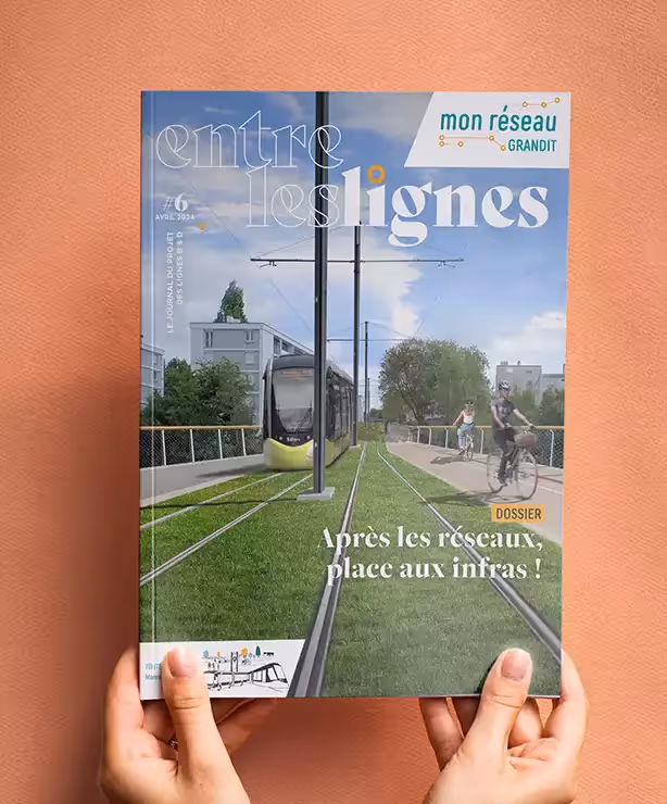 Une personne tient le magazine "Entre les lignes" devant un fond orange. La couverture présente un tramway roulant au centre d'une piste cyclable avec le texte "Après les réseaux, place aux infras !". Titre : Lecture du magazine "Entre les lignes"