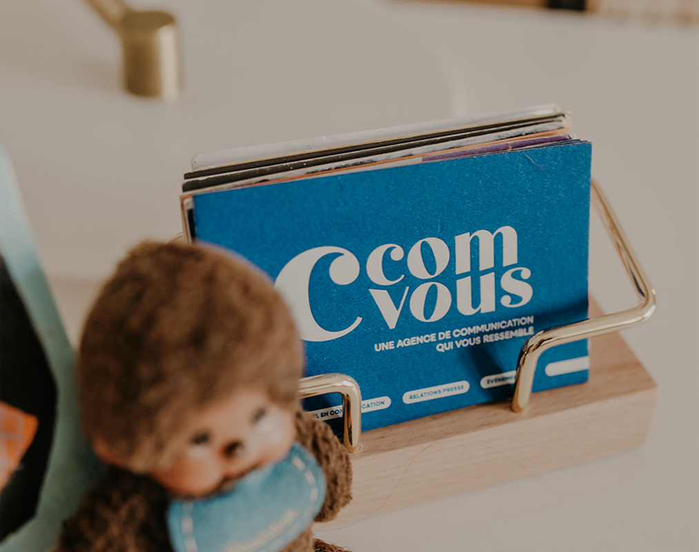 Cartes de visite C’ComVous dans un porte-cartes en bois et laiton, avec logo bleu et slogan imprimé.