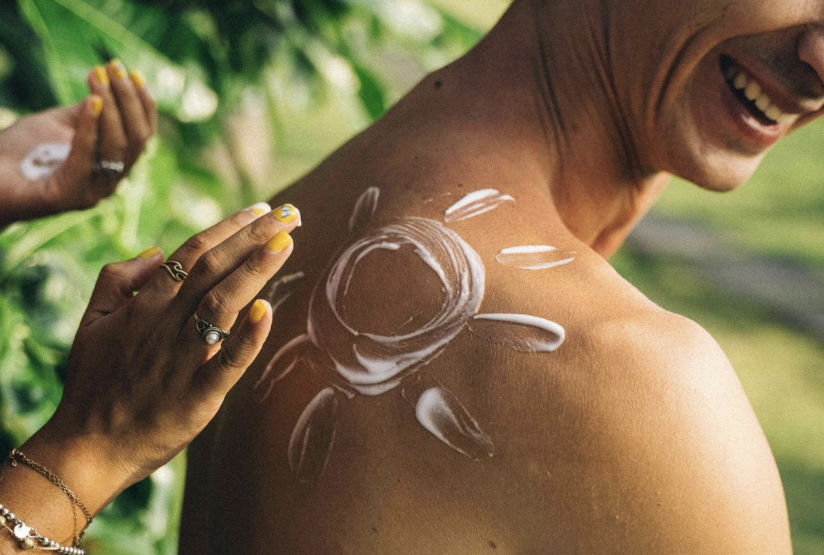 Person trägt Sonnencreme in Form einer Sonne auf den Rücken eines lachenden Mannes auf – Symbol für Sonnenschutz und Hautpflege im Sommer.