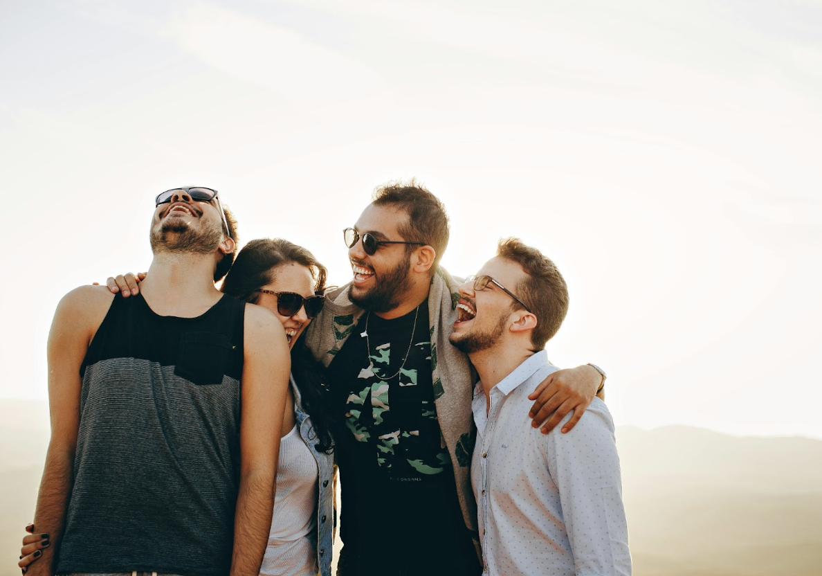 Vier junge Erwachsene stehen eng beieinander, lachen herzlich und genießen einen sonnigen Moment im Freien.