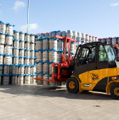 Yellow forklift moving metal kegs stacked outdoors under a blue sky.