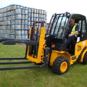 Forklift operator handling a yellow forklift with extended forks outside on grass near stacked pallets wrapped in plastic.