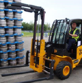 Forklift operator moving stacked keg barrels outdoors on a paved surface.