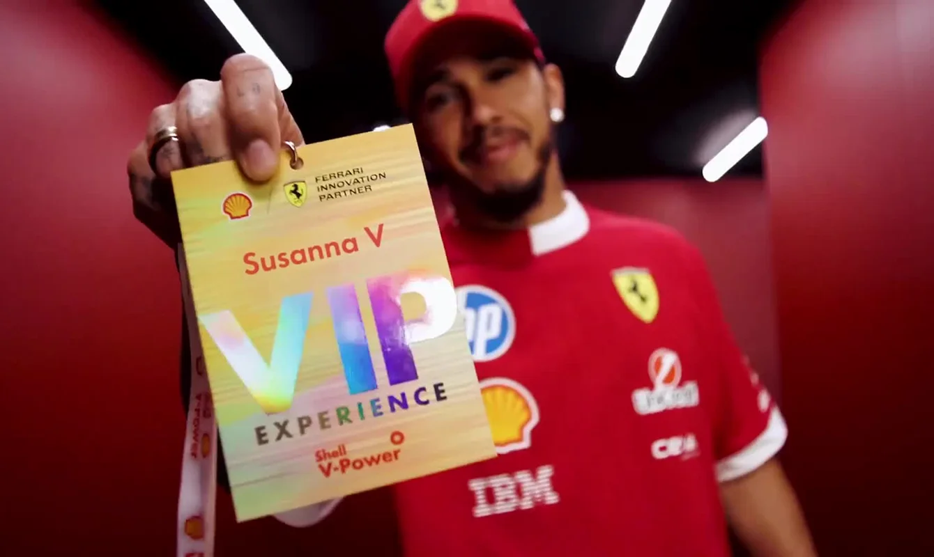 Lewis Hamilton in F1 uniform holding a VIP Experience pass labeled Susanna V with logos of Shell and Ferrari Innovation Partner.