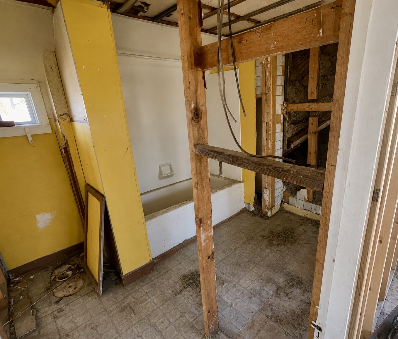 The upstairs bathroom mid-demolition: yellow walls, an old tub, and the ceiling opened to the rafters