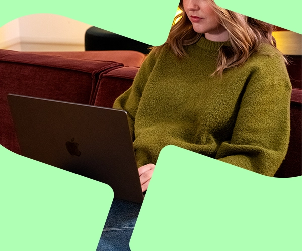 Kvinna i en grön tröja som arbetar på en bärbar dator på en vinröd soffa mot en grön bakgrund.
